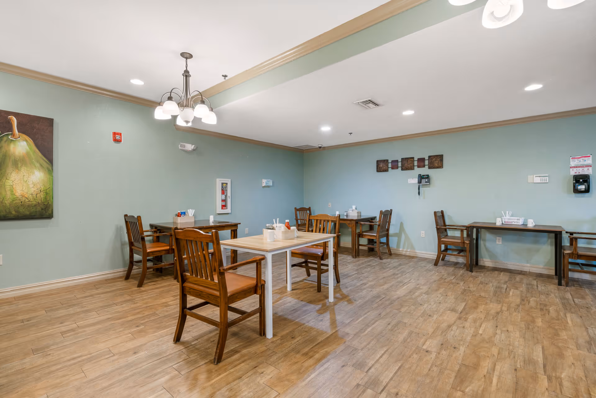 A dining area with several wooden tables and chairs arranged on a wood-patterned floor. The walls are painted light green and decorated with a large painting of a pear and a set of small square wall art. The ceiling has recessed lighting and a chandelier with multiple lights. Various items like napkin holders and condiments are placed on the tables.