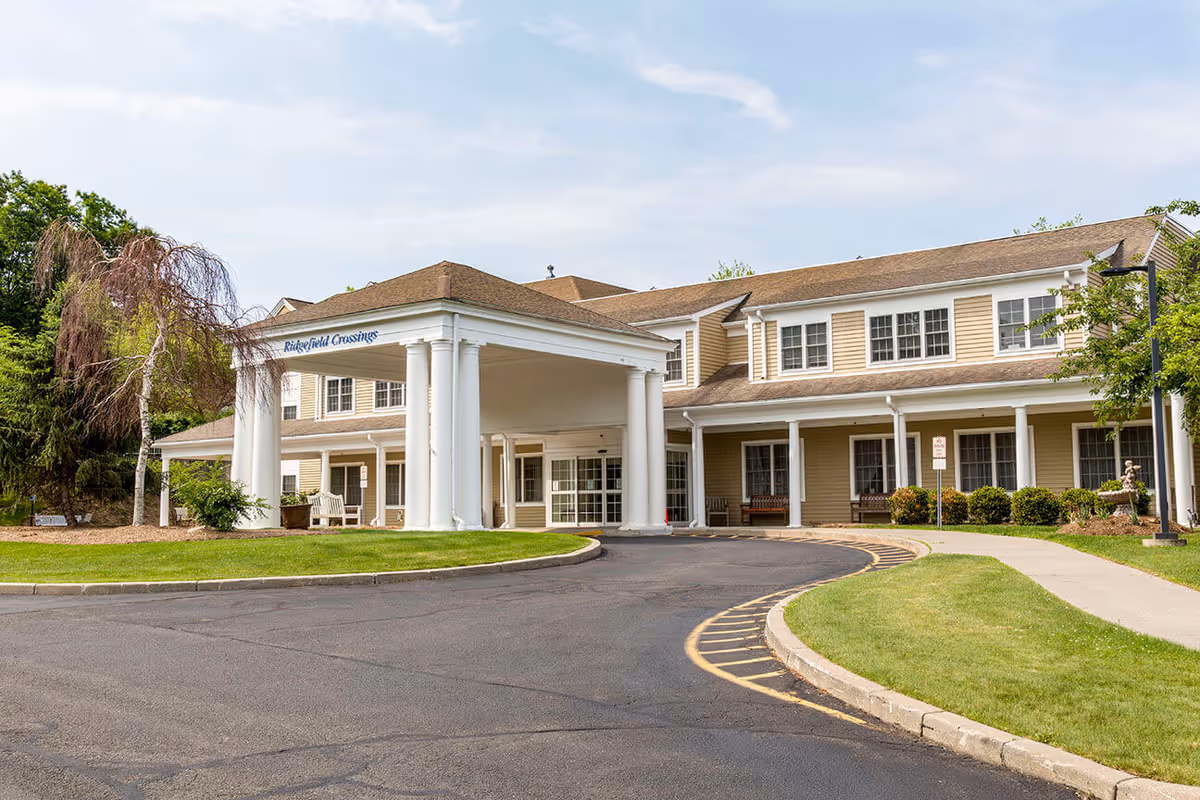 Exterior view of Benchmark at Ridgefield Crossings senior living facility showing a two-story building with beige siding, white columns, and a covered entrance driveway. The surrounding area includes a curved asphalt driveway, green lawn, trees, and benches near the entrance.