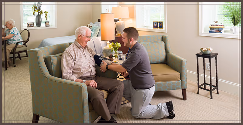 An elderly man sitting on a patterned armchair in a well-lit living room area while a healthcare professional kneels in front of him, checking his blood pressure with a cuff. The room has large windows, a small table with books and a plant, and another elderly person sitting at a table in the background.