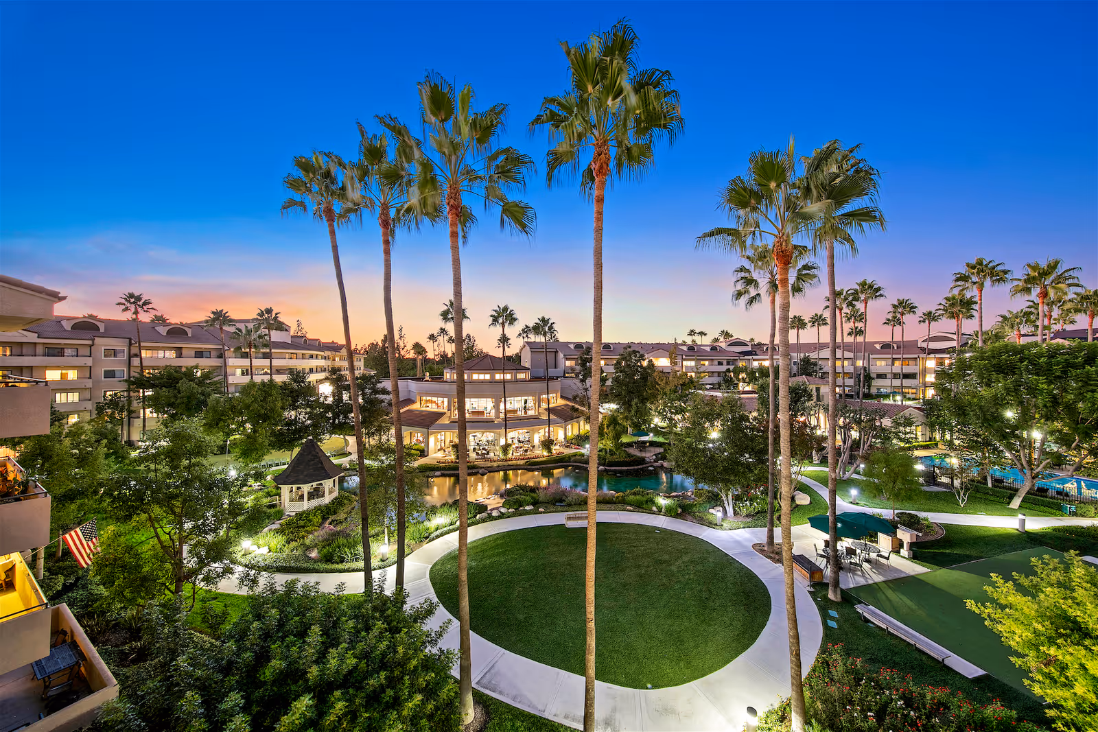 Well-lit senior living community courtyard at dusk with tall palm trees, a circular lawn, gazebo, pond, and surrounding multi-story buildings.