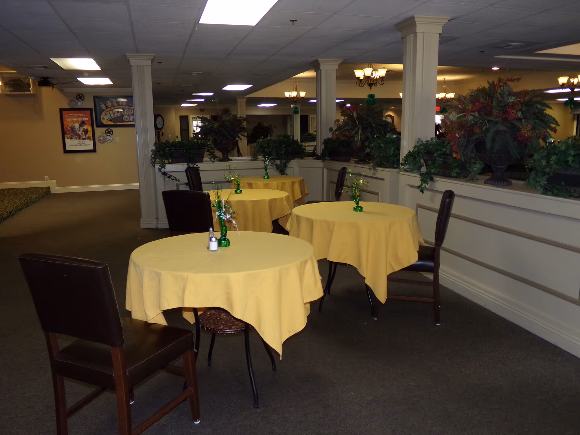 Small dining area with round tables covered in yellow tablecloths, chairs, and potted plants along a half wall.