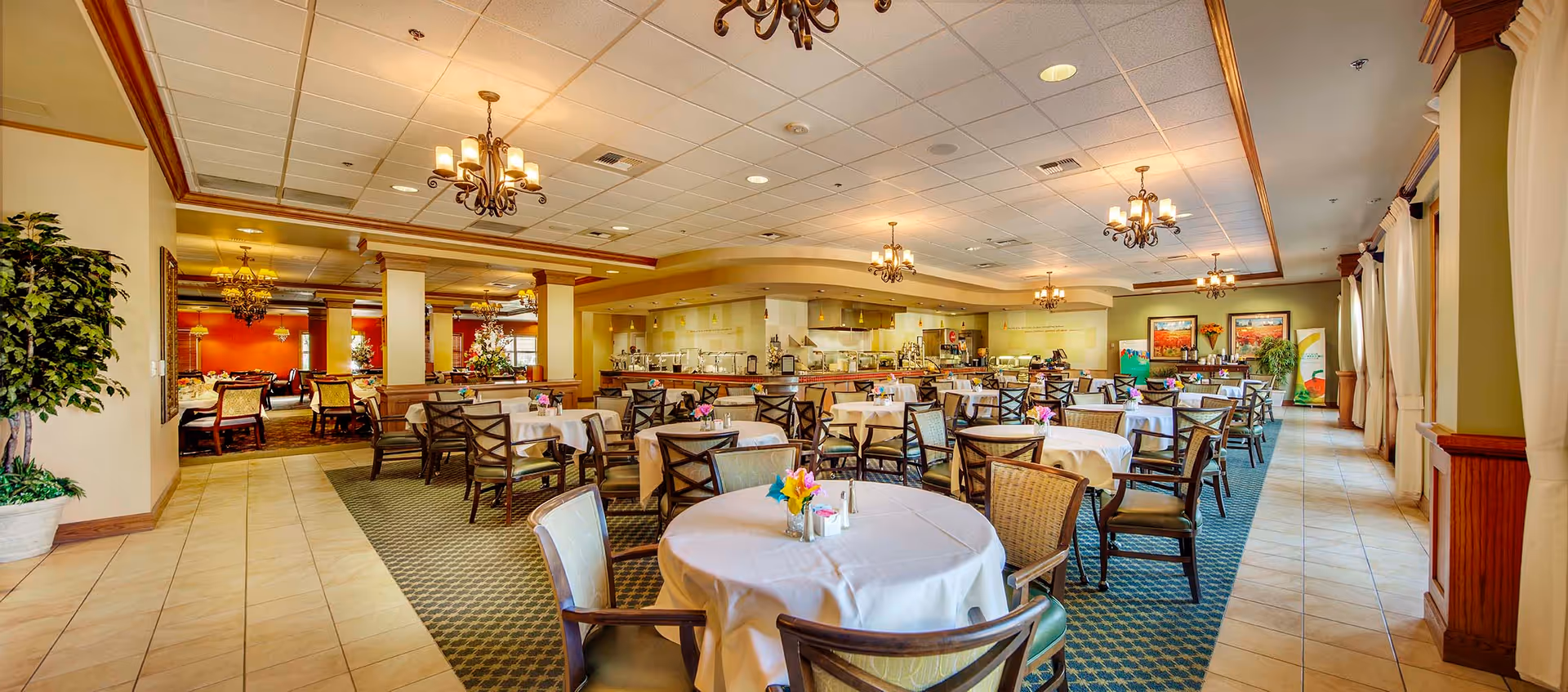Spacious dining room with round tables draped in white cloths, wooden chairs, chandeliers, and a serving area in the background.
