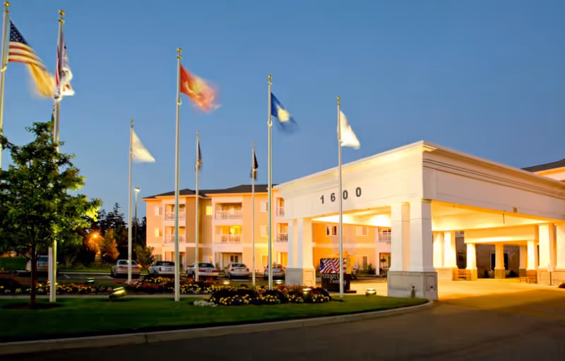 Exterior view of Patriot's Landing senior living facility at dusk with multiple flagpoles displaying flags in front of the building entrance marked with the number 1600. The building is illuminated with warm lights and surrounded by landscaped greenery and flowers.