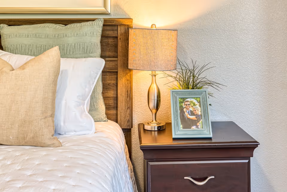 Bedside view showing a made bed with pillows and a wooden headboard, and a nightstand with a lamp and framed photo.