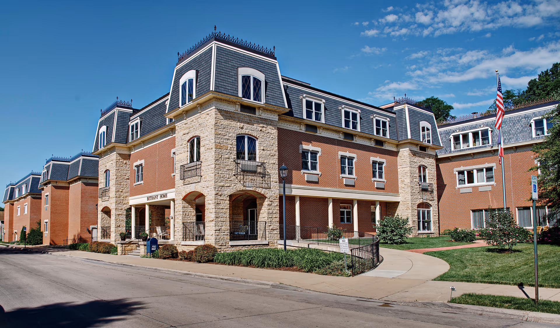Exterior view of Bethany Home Retirement Center, a large brick building with stone accents and multiple windows. The building has a mansard roof with dormer windows and decorative ironwork on top. There is a sidewalk and a small landscaped area with grass and bushes in front, along with an American flag on a flagpole.