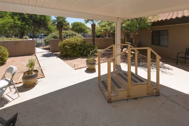 Outdoor covered patio area with a wooden ramp, potted plants, folding chairs, and a pathway leading to a gated entrance surrounded by bushes and palm trees.