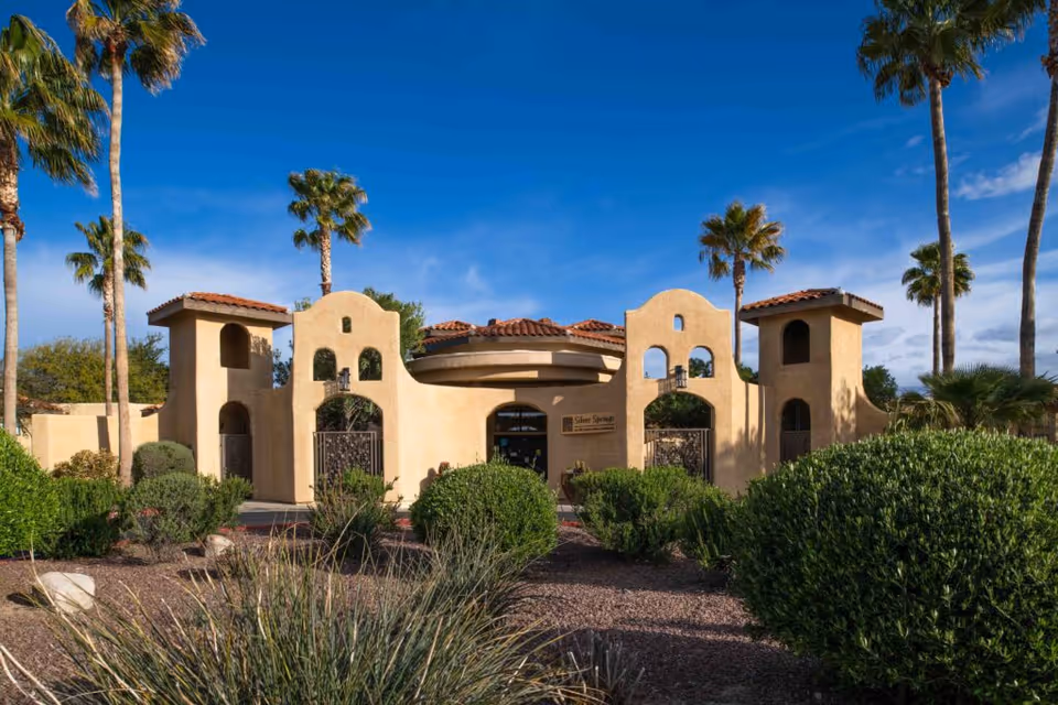 Exterior view of Silver Springs facility with a southwestern architectural style featuring beige stucco walls, arched openings, and red tile roofs. The entrance is framed by two towers and surrounded by desert landscaping with bushes, palm trees, and gravel under a clear blue sky.