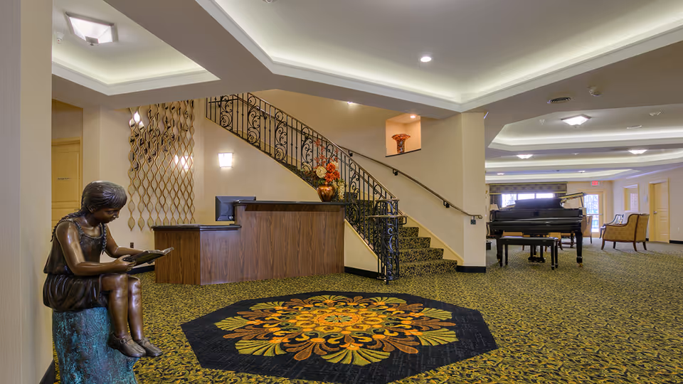 Interior view of a senior living facility lobby with a decorative carpet, a bronze statue of a girl reading a book, a wooden reception desk, a staircase with ornate railing, a grand piano, and seating area with chairs. The space is well-lit with ceiling lights and has a warm, welcoming atmosphere.