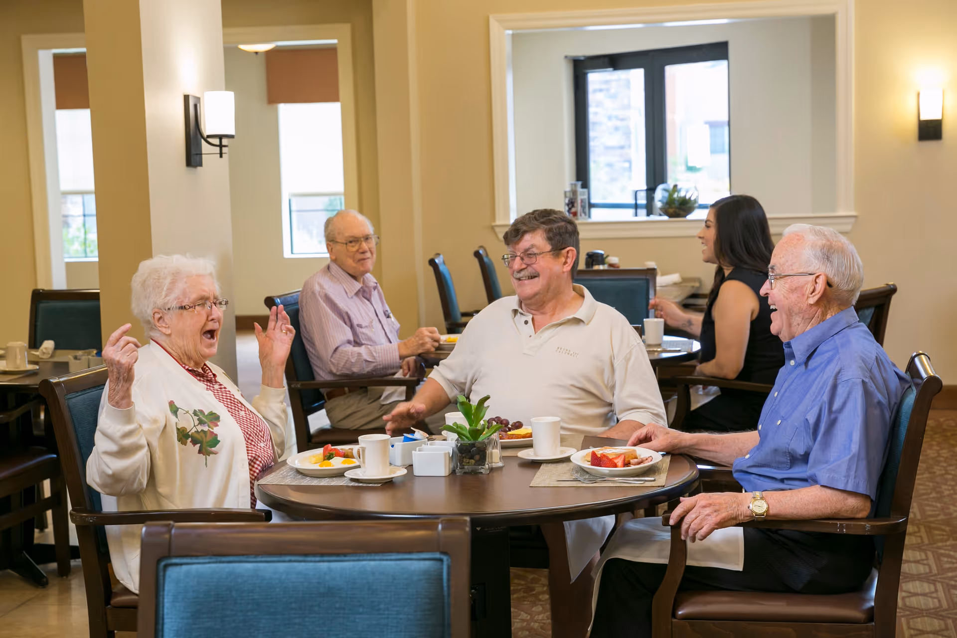 A group of older adults and a younger woman smiling and dining around a round table in a bright senior living dining room.