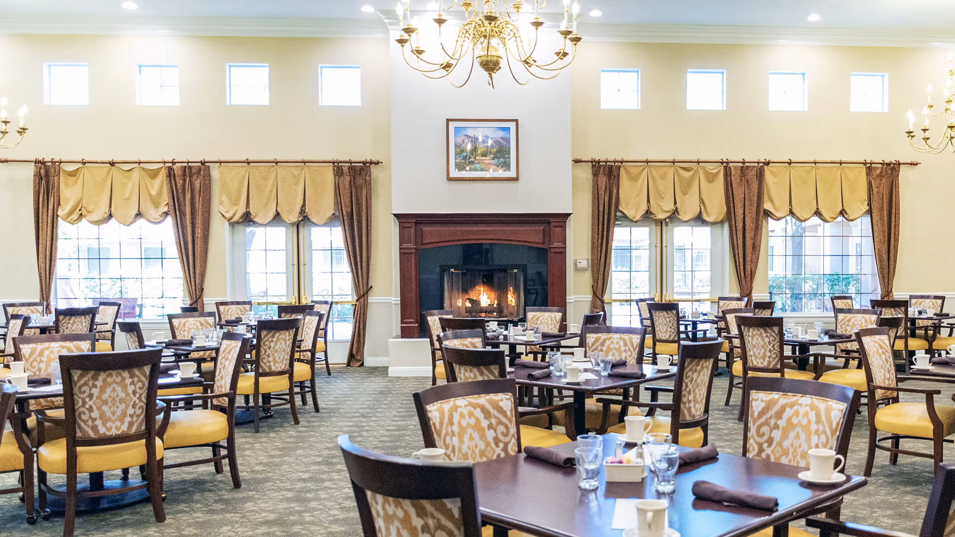 Spacious dining room with multiple set tables and upholstered chairs, chandeliers and a central fireplace.