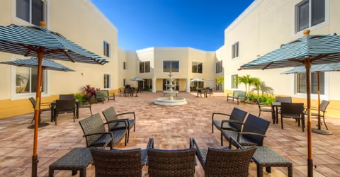 Outdoor courtyard area at Discovery Village At The Forum with multiple seating arrangements including wicker chairs and tables shaded by striped umbrellas, surrounded by a two-story building with windows, and a central water fountain under a clear blue sky.