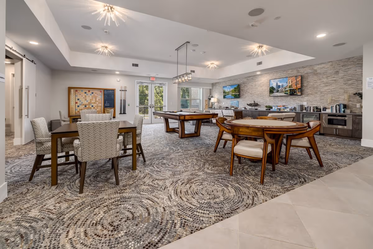Bright communal activity and dining room with tables, chairs, a pool table, TVs, and a kitchenette.