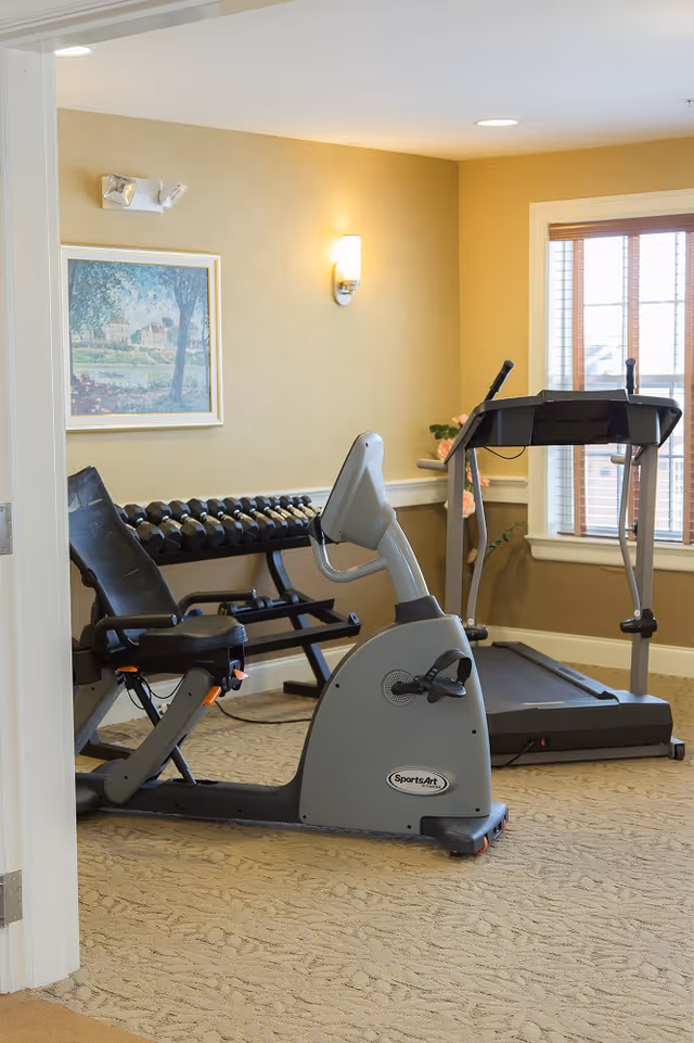 A small fitness room with exercise equipment including a recumbent stationary bike, a treadmill, and a rack of dumbbells. The room has beige walls, a window with blinds, a framed painting on the wall, and a wall-mounted light fixture.