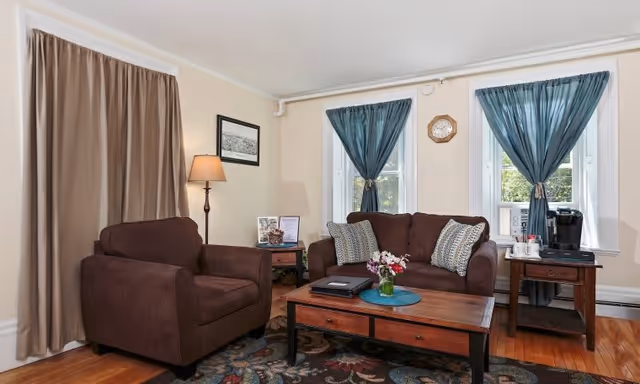 A cozy living room with a brown armchair and a matching brown loveseat adorned with patterned pillows. There is a wooden coffee table with a vase of flowers and a laptop on it. Two windows with blue curtains are behind the loveseat, and a small side table with a coffee maker and cups is next to it. A floor lamp and framed artwork are on the wall beside the armchair. The room has hardwood floors and a patterned area rug.