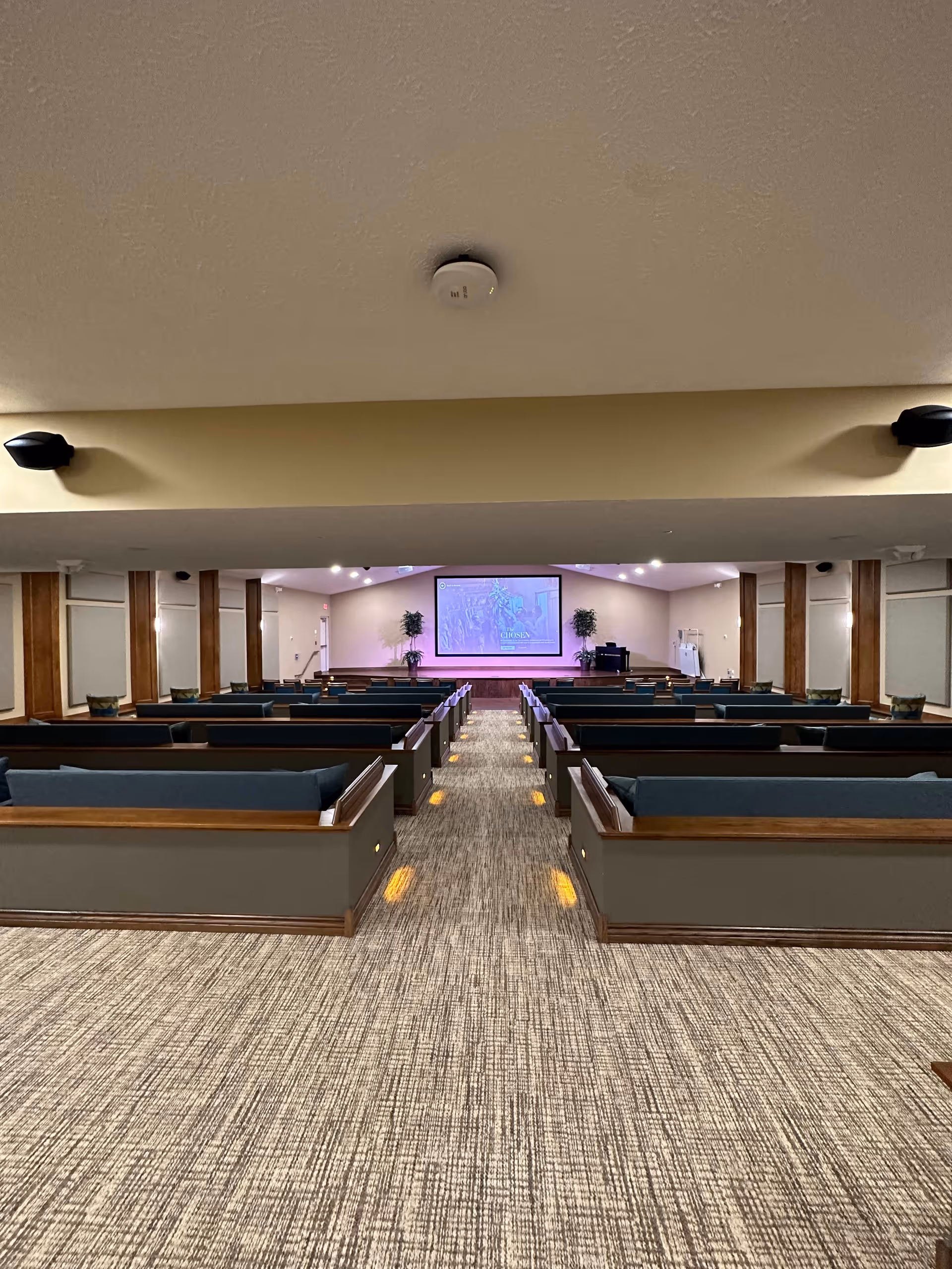 A large room with rows of wooden pew-style seating with blue cushions facing a stage with a large screen displaying an image. The room has beige walls, carpeted floor, and ceiling lights. There are two potted plants on either side of the stage and a podium on the right side.