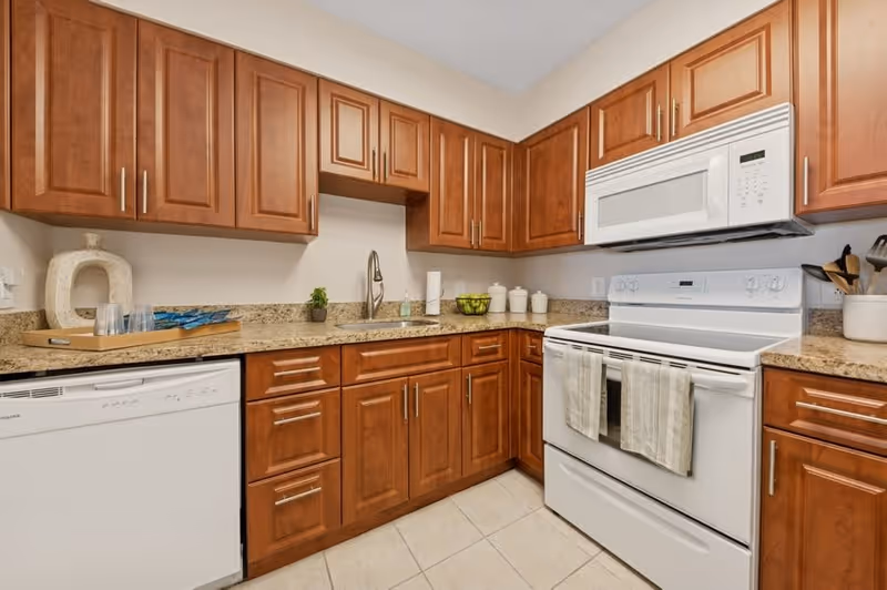A kitchen with wooden cabinets, granite countertops, a white dishwasher, a white stove with a microwave above it, and various kitchen items including a tray with glasses, a small plant, paper towels, a bowl of green apples, and ceramic containers.