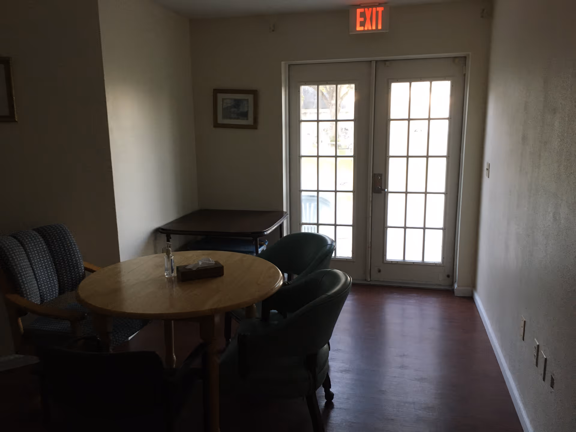 A small room with a round wooden table surrounded by four chairs, two upholstered and two green cushioned. There is a small table against the wall and a framed picture hanging above it. Double glass doors with multiple panes lead outside, with an illuminated exit sign above the doors. The floor is wooden and the walls are plain.