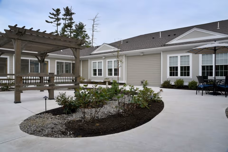 Outdoor courtyard area of a senior living facility featuring a wooden pergola with benches, a landscaped garden bed with small plants and shrubs, concrete walkways, and a seating area with a table and umbrella. The building exterior is light gray with white trim and multiple windows.