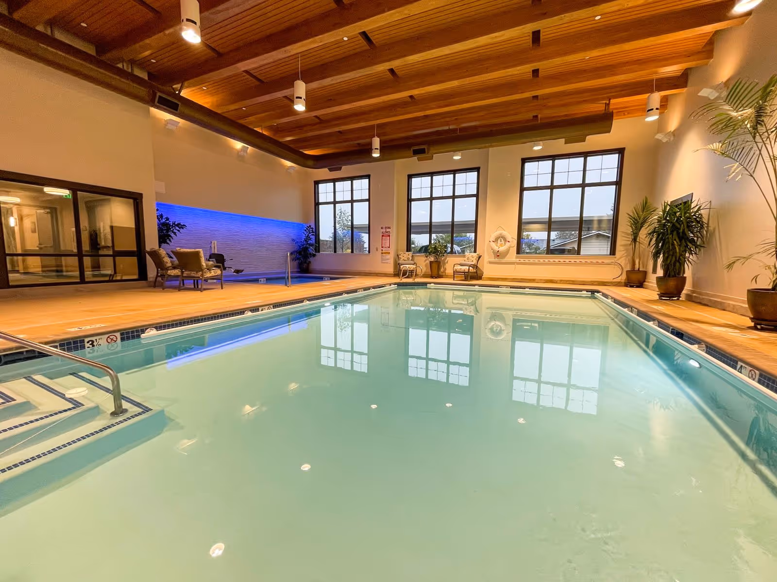 Indoor swimming pool area with clear water, steps leading into the pool, large windows letting in natural light, potted plants, chairs, and a wooden ceiling with hanging lights.