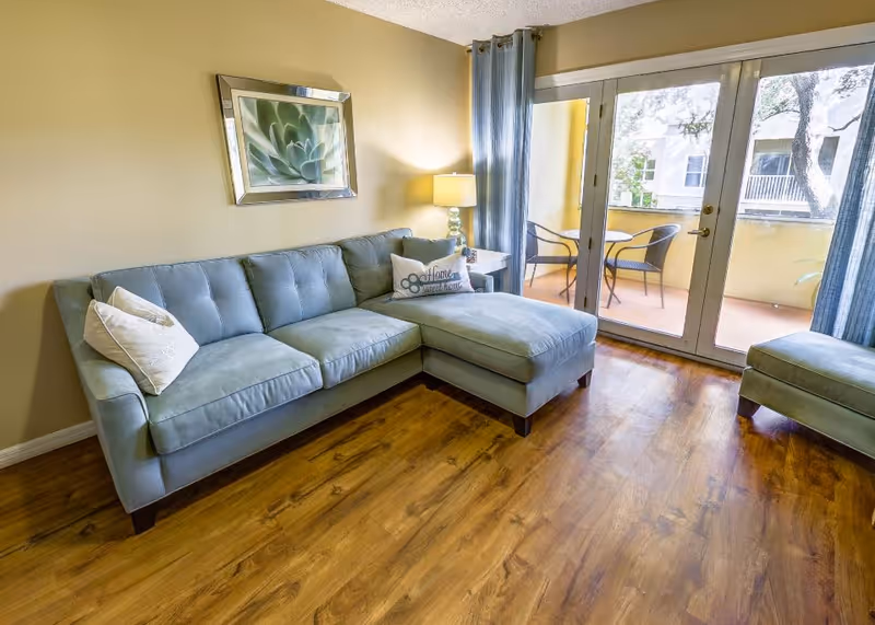 A cozy living room with a light blue sectional sofa adorned with decorative pillows, a side table with a lamp, a framed succulent artwork on the beige wall, and large glass doors leading to a balcony with two chairs and a small table.