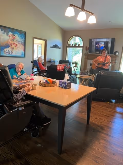 Seniors seated in a living-room style common area around a table while a person plays guitar near a TV and fireplace.