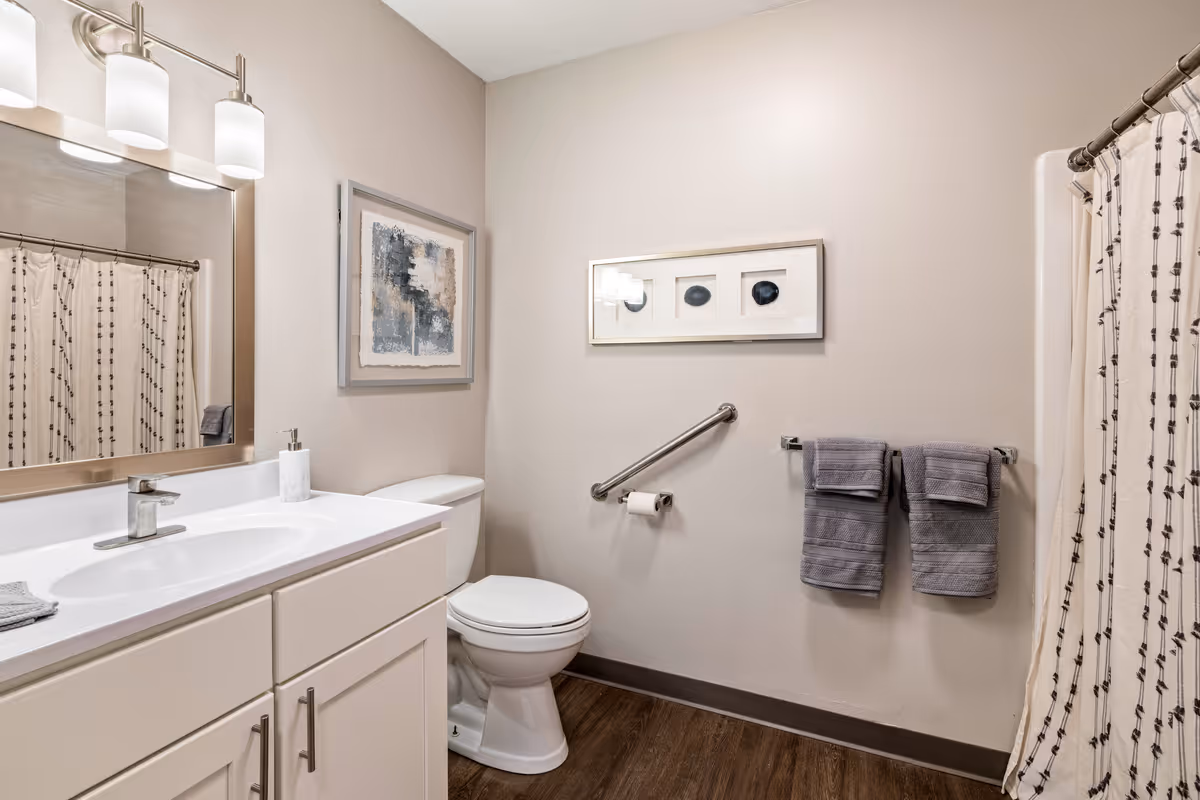 A clean and modern bathroom featuring a white vanity with a sink and faucet, a large mirror with three light fixtures above it, a toilet with a safety grab bar next to it, two gray towels hanging on a towel rack, a shower with a patterned curtain, and two framed abstract artworks on the walls. The floor is wood-style and the walls are painted light beige.