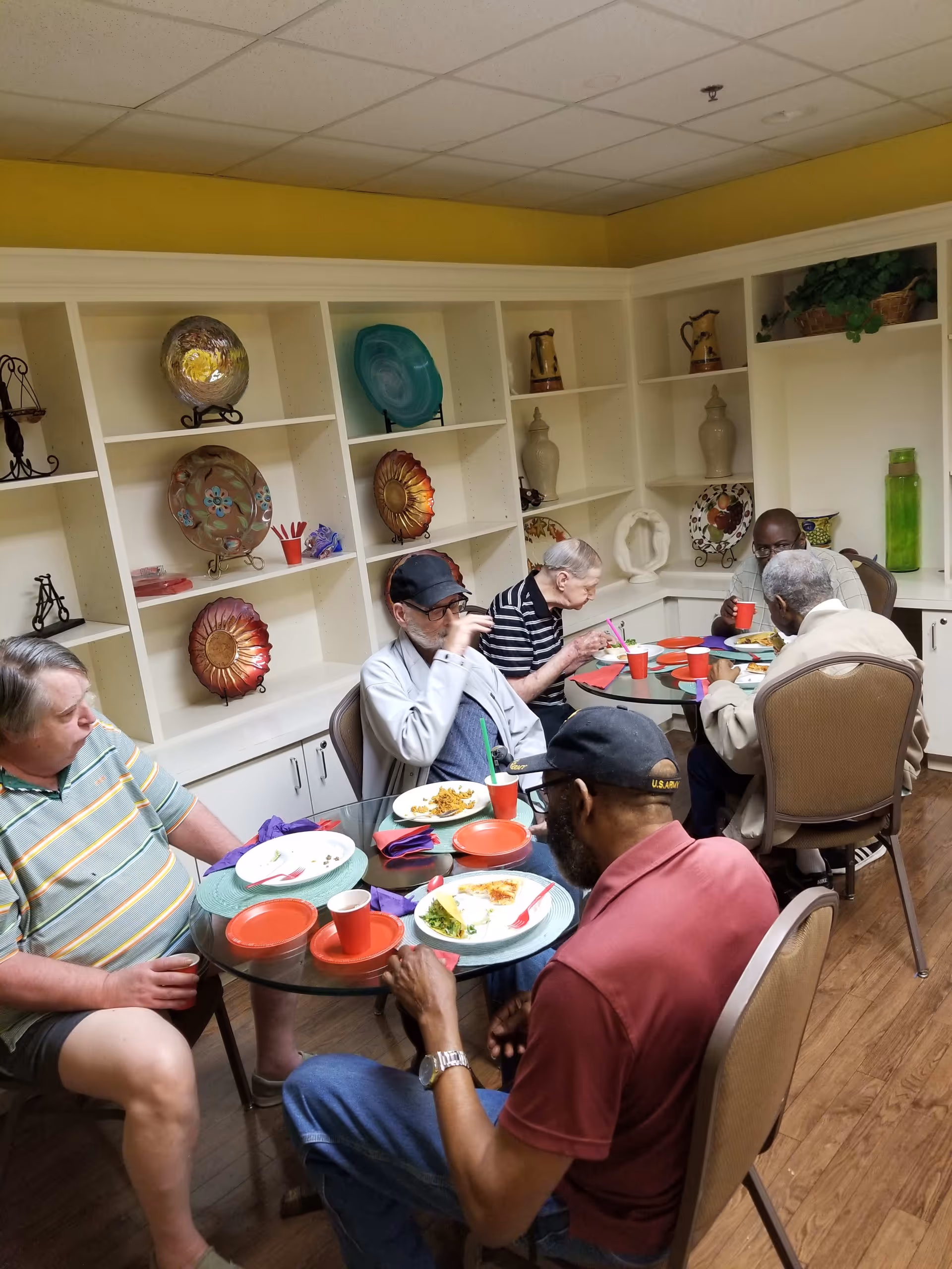 Five elderly men sitting around a glass dining table in a room with white shelves displaying decorative plates and vases, eating a meal and drinking from red cups.