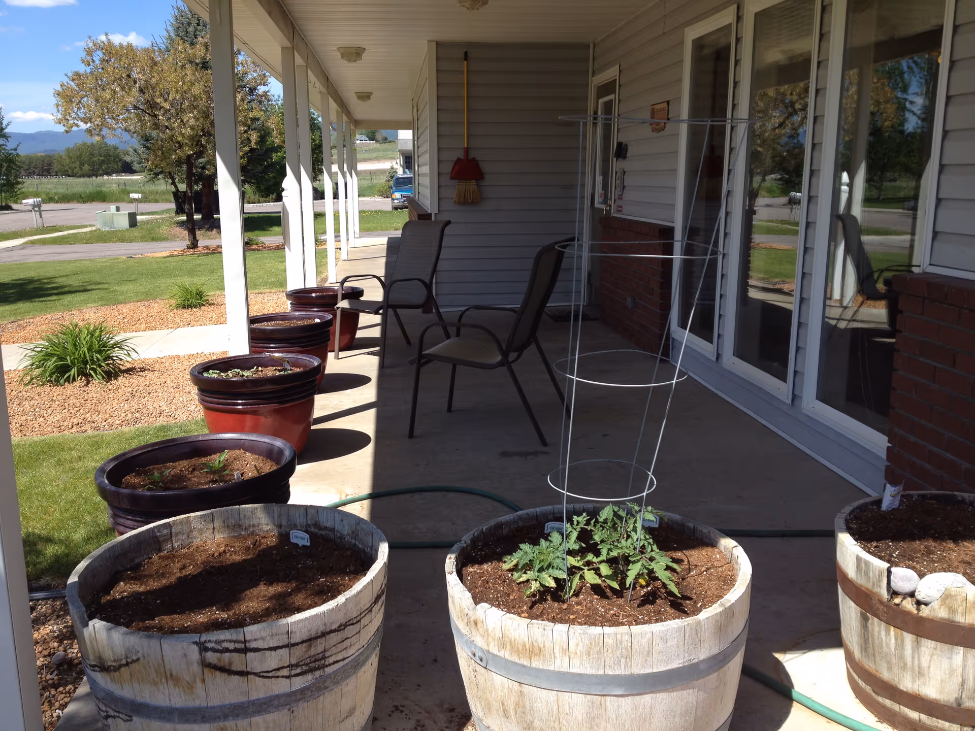 A covered outdoor patio area with several large planters containing soil and some small plants. There are two chairs and a broom hanging on the wall. The patio overlooks a grassy area with trees and a road in the background.