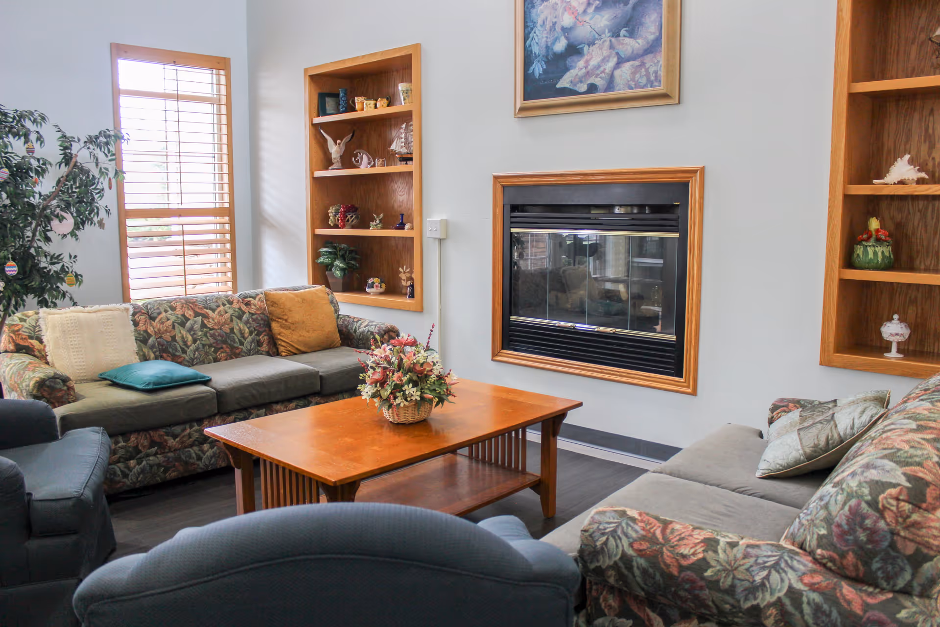 A cozy living room with floral patterned sofas, a wooden coffee table with a flower basket centerpiece, built-in wooden shelves with decorative items, a fireplace framed in wood, and a window with wooden blinds letting in natural light.