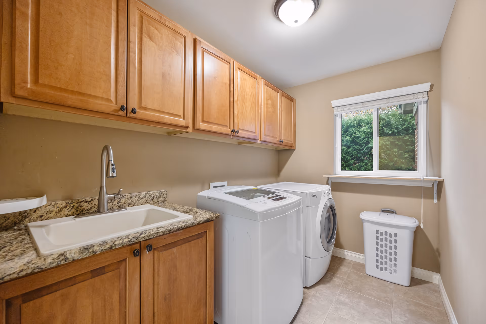 Laundry room with a white top-loading washing machine and a front-loading dryer side by side. Above them are wooden cabinets with dark knobs. There is a granite countertop with a built-in sink and a modern faucet on the left side. A window with white blinds lets in natural light and shows greenery outside. A white laundry basket is placed on the floor near the window.