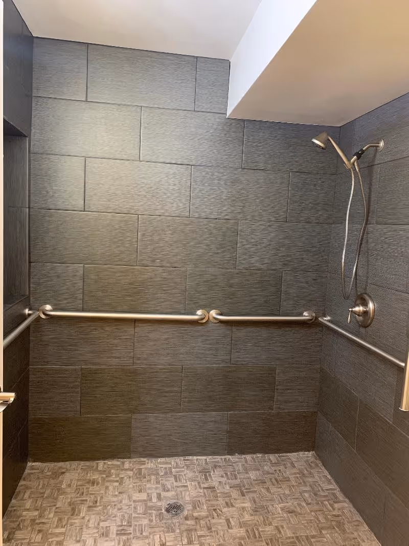 A tiled shower area with dark gray rectangular wall tiles and a mosaic-patterned floor. The shower features a handheld showerhead attached to a flexible hose, and there are multiple horizontal stainless steel grab bars installed on the walls for safety.