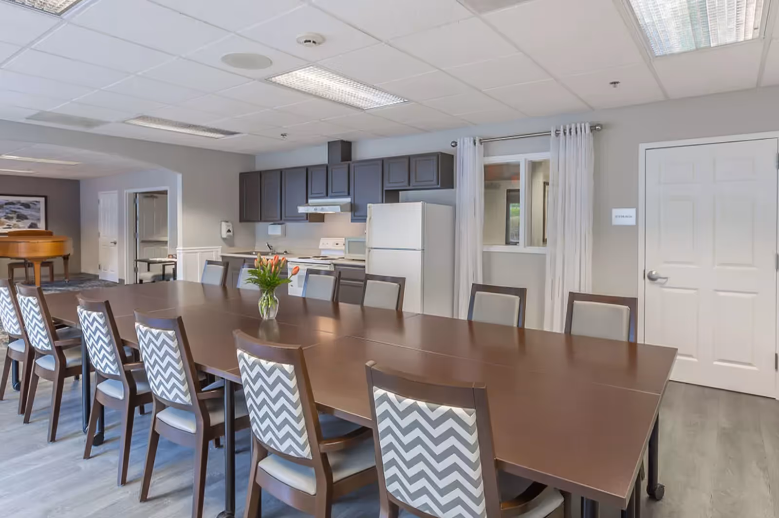 Communal dining room with a long wood table, patterned chairs, a vase of flowers, and a kitchenette in the background.