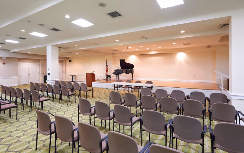 A large room set up for a presentation or performance with rows of chairs facing a small stage. On the stage, there is a grand piano, a podium, and an American flag. The room has a patterned green carpet and white walls with recessed lighting in the ceiling.