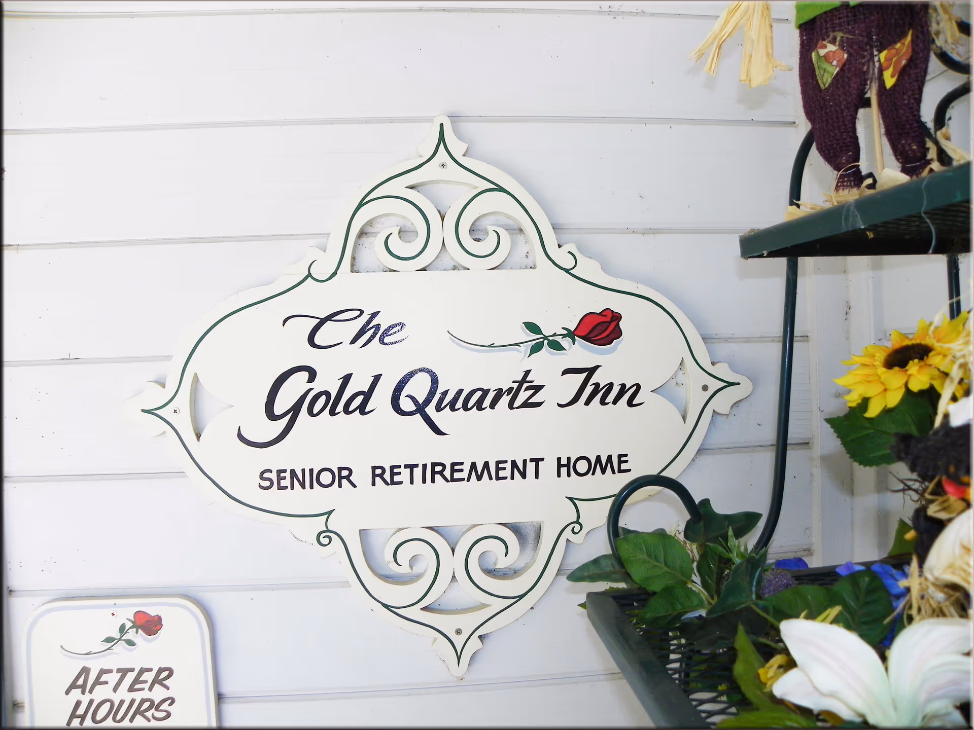 Decorative sign reading "The Gold Quartz Inn SENIOR RETIREMENT HOME" mounted on a white paneled wall with potted plants nearby.