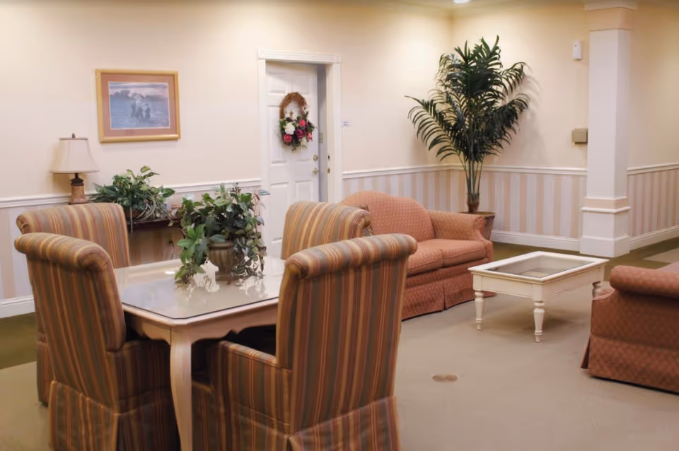 A cozy living room area with a small square table surrounded by four striped upholstered chairs. There are two peach-colored sofas, a white coffee table, a tall potted plant in the corner, a side table with a lamp and plants, and a white door decorated with a floral wreath. The walls are light-colored with a striped wallpaper border.