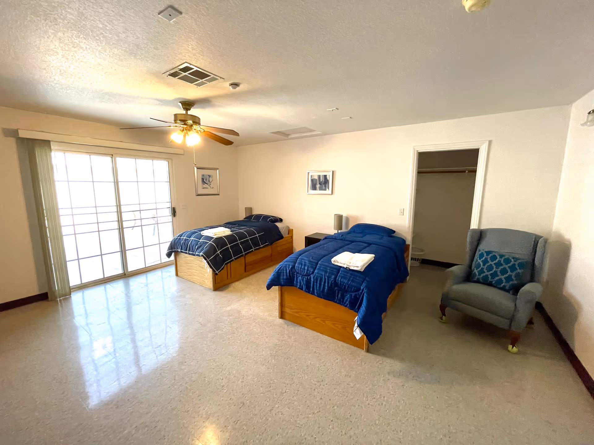 Spacious bedroom with two twin beds, a blue armchair, sliding glass doors and a ceiling fan.