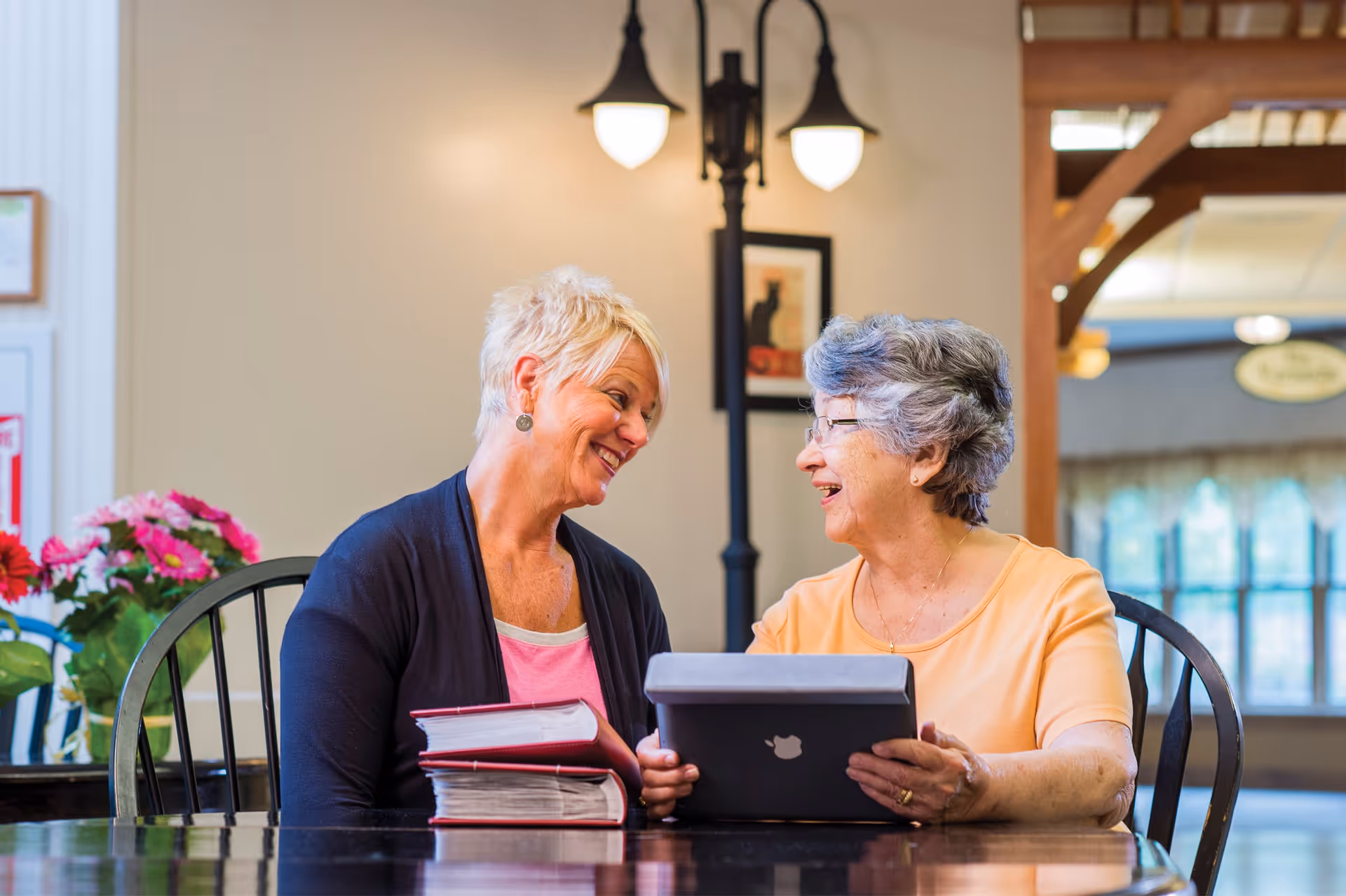 Two elderly women sitting at a table indoors, smiling and looking at each other. One woman holds a tablet device while the other has two thick binders stacked on the table. There are flowers and a lamp post-style light fixture in the background.