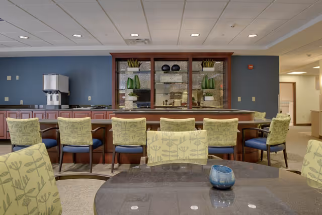 Interior view of a senior living facility dining area with a row of chairs lined up against a counter. The chairs have light green upholstery with a plant pattern and dark wooden legs. Behind the counter is a wooden cabinet with glass shelves displaying decorative plants and plates. In the foreground, there is a round table with a small blue decorative item on it. The walls are painted in blue and beige tones, and the ceiling has recessed lighting.