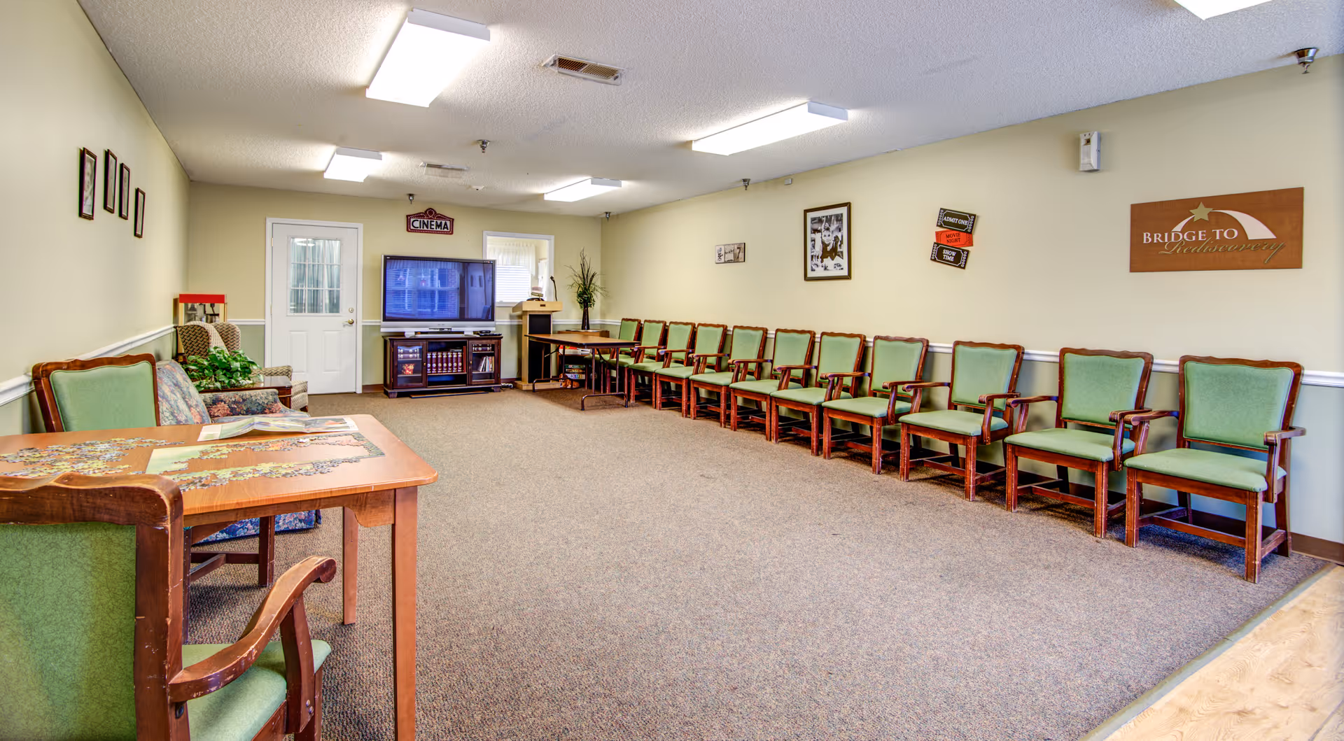 A spacious room with a long row of green cushioned wooden chairs lined up against a beige wall. There is a table with a puzzle in progress and a few chairs in the foreground. At the far end of the room, there is a TV on a stand with a sign above it that reads 'CINEMA'. The walls are decorated with framed pictures and a sign that says 'BRIDGE TO Independence'. The room has a carpeted floor and fluorescent ceiling lights.