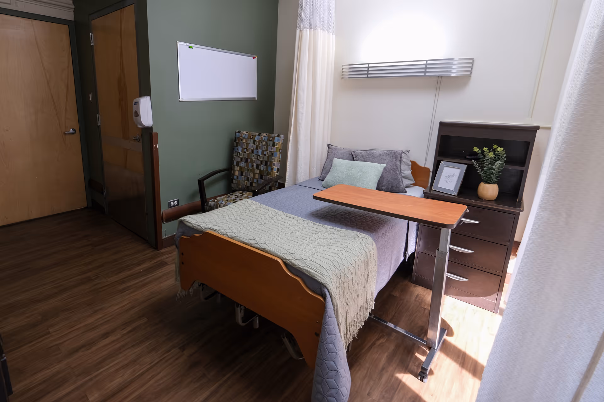 A small, neatly arranged bedroom in a senior living facility with a single bed covered in gray and green bedding. There is a wooden overbed table positioned over the bed, a patterned armchair next to the bed, and a dark wooden dresser with a potted plant and framed picture on top. The room has wood flooring, a green accent wall, and a whiteboard mounted on the wall.