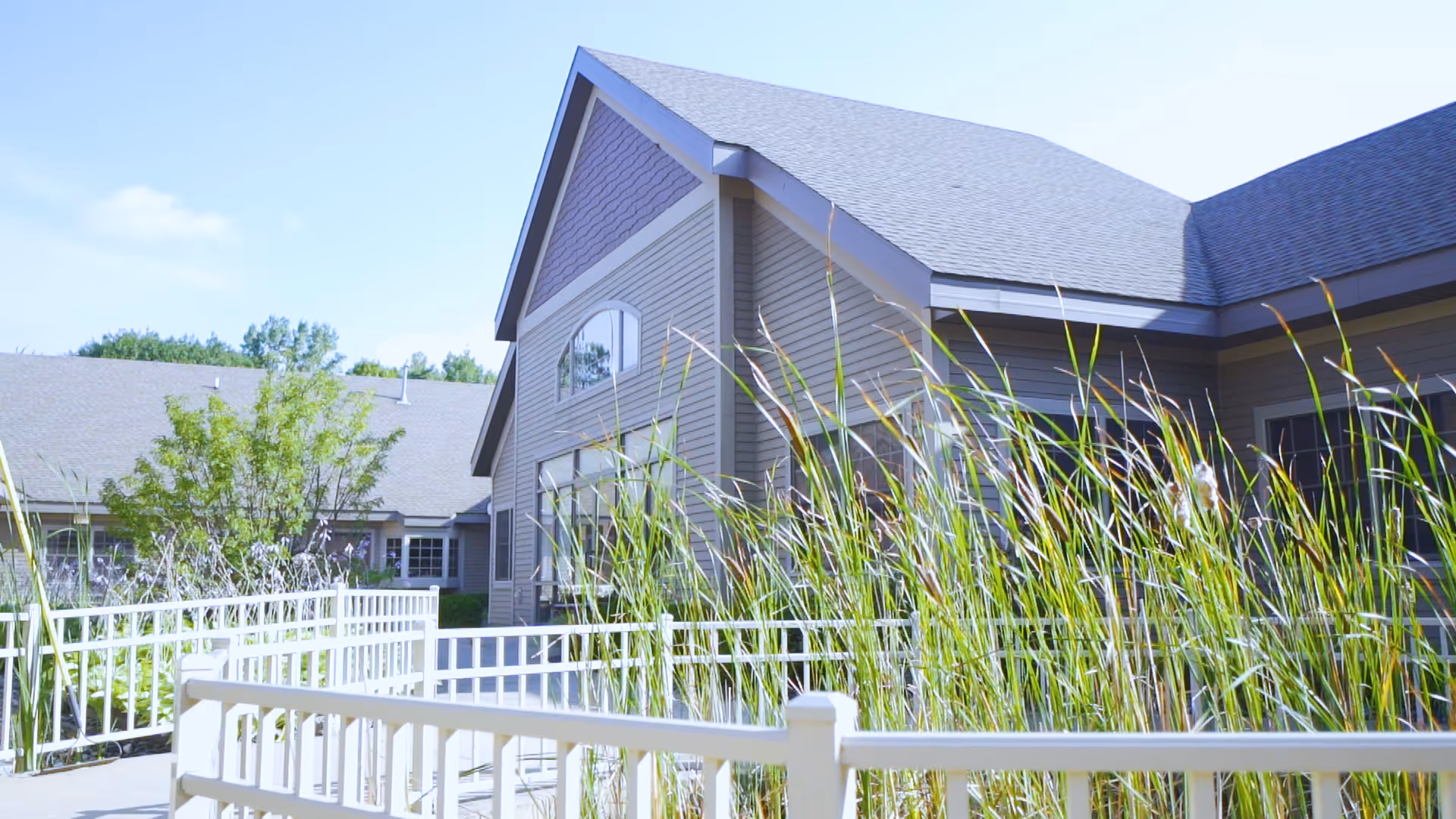 Exterior view of a senior living facility building with beige siding and a gray roof, surrounded by tall grasses and a white fence under a clear blue sky.
