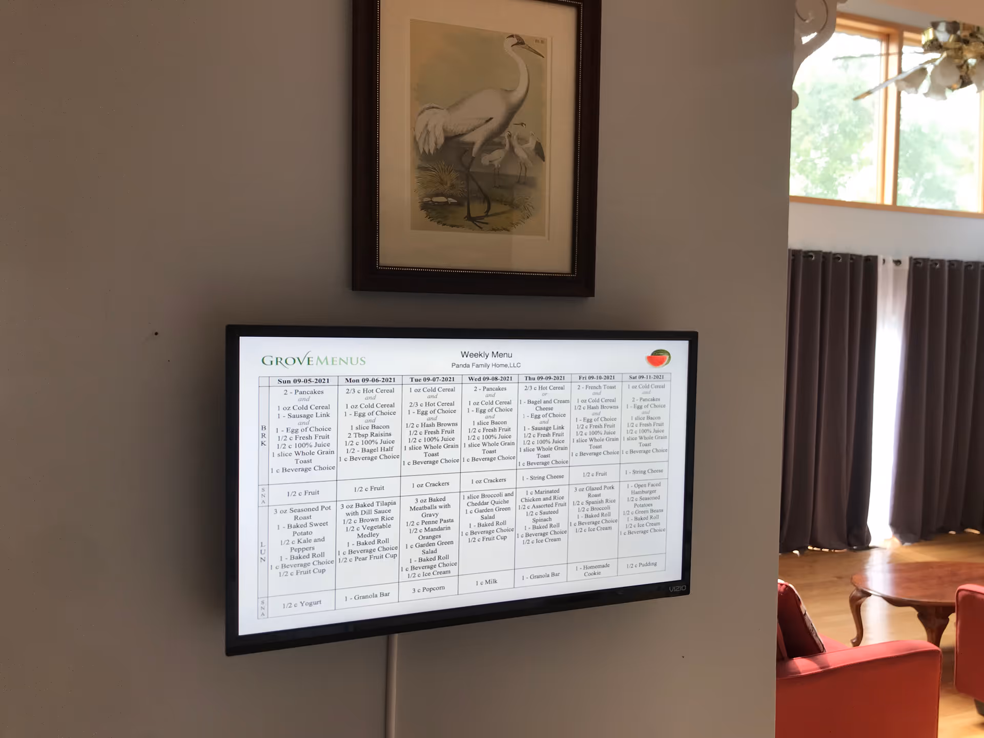 A wall-mounted TV screen displaying a weekly menu for Panda Family Home, LLC, with a framed picture of a white bird above it. To the right, part of a living room is visible with large windows, curtains, and red furniture.