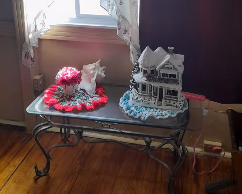 A glass-top metal table positioned in front of a window with floral curtains. On the table, there is a small white vase with pink flowers on a red and white crocheted doily, a large white seashell, and a decorative miniature house covered in snow on a blue and white crocheted doily. The floor is wooden, and a pink electrical cord is plugged into an outlet near the table.