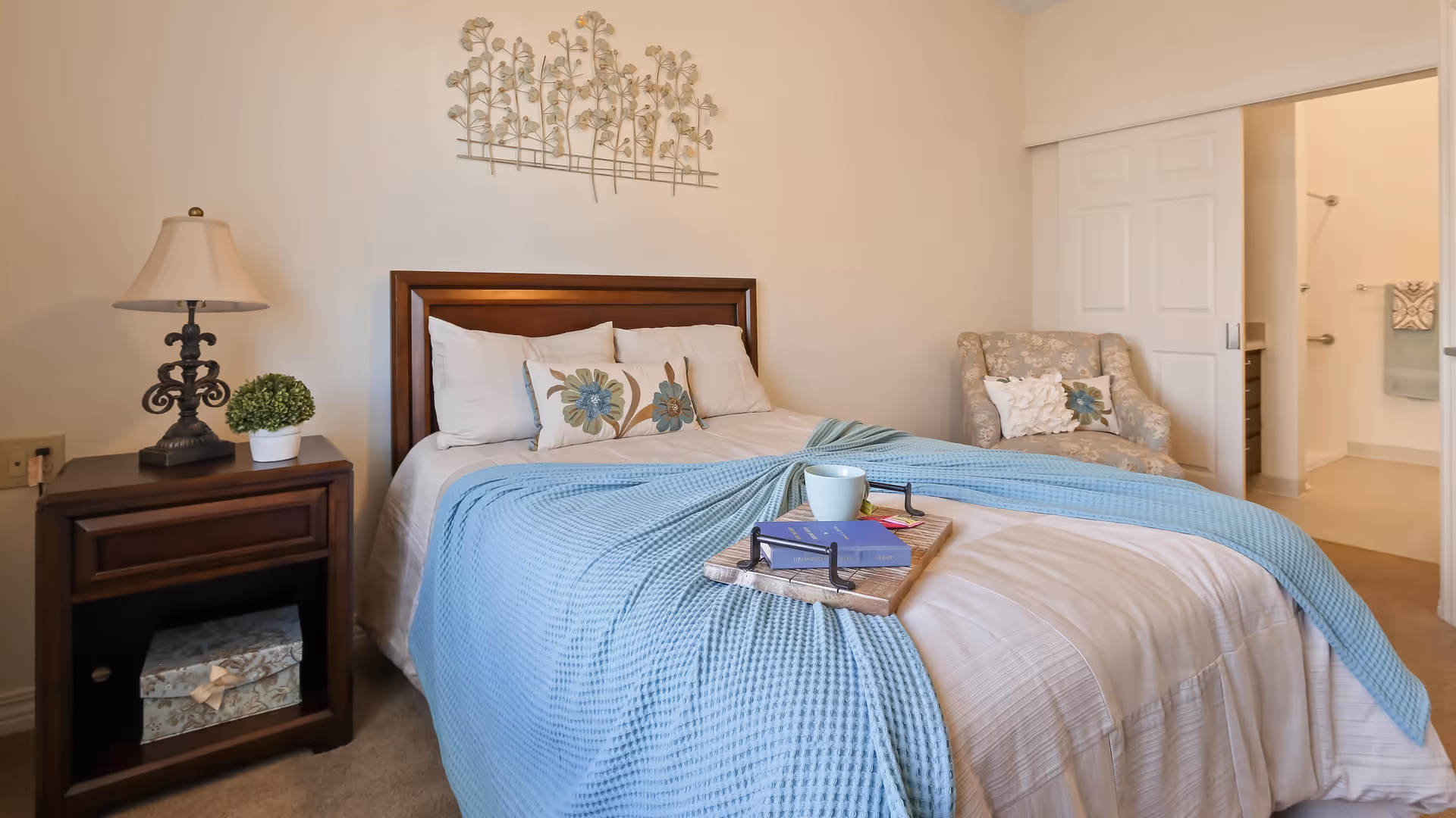 A cozy bedroom with a neatly made bed featuring white and blue bedding, decorative pillows with floral designs, and a wooden headboard. A wooden nightstand with a lamp and a small potted plant is beside the bed. There is a cushioned armchair with floral upholstery and pillows in the corner near a doorway leading to a bathroom.