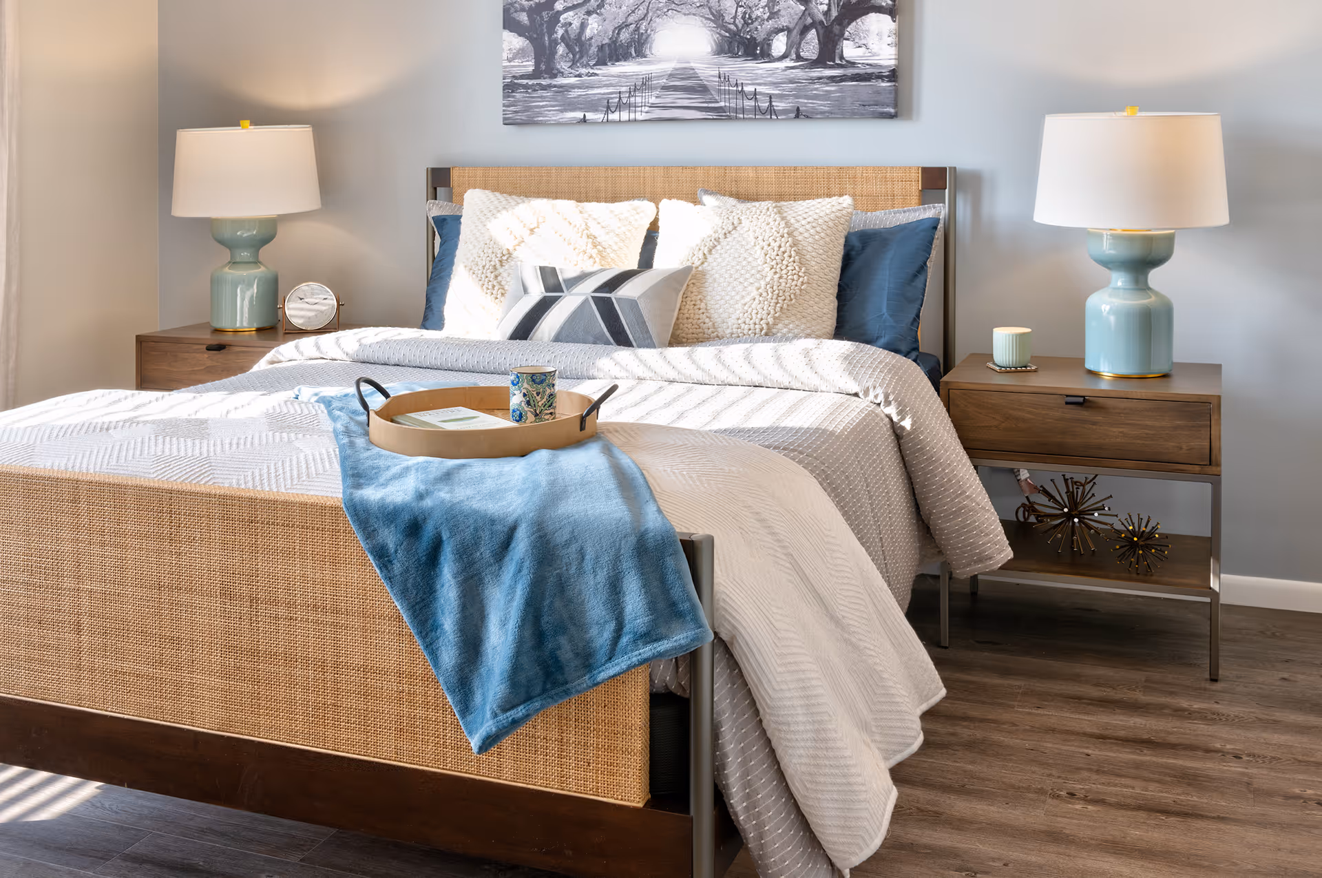 A neatly made bed with a woven headboard and footboard, adorned with multiple pillows in white and blue. A blue throw blanket and a wooden tray with a cup and a book rest on the bed. On each side of the bed are wooden nightstands with light blue ceramic lamps and decorative items. Above the bed hangs a black and white photograph of a tree-lined path.