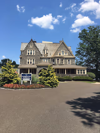 Victorian-style multi-story stone building with a sign reading 'Langhorne Gardens' in front, a circular driveway, and trees under a blue sky.