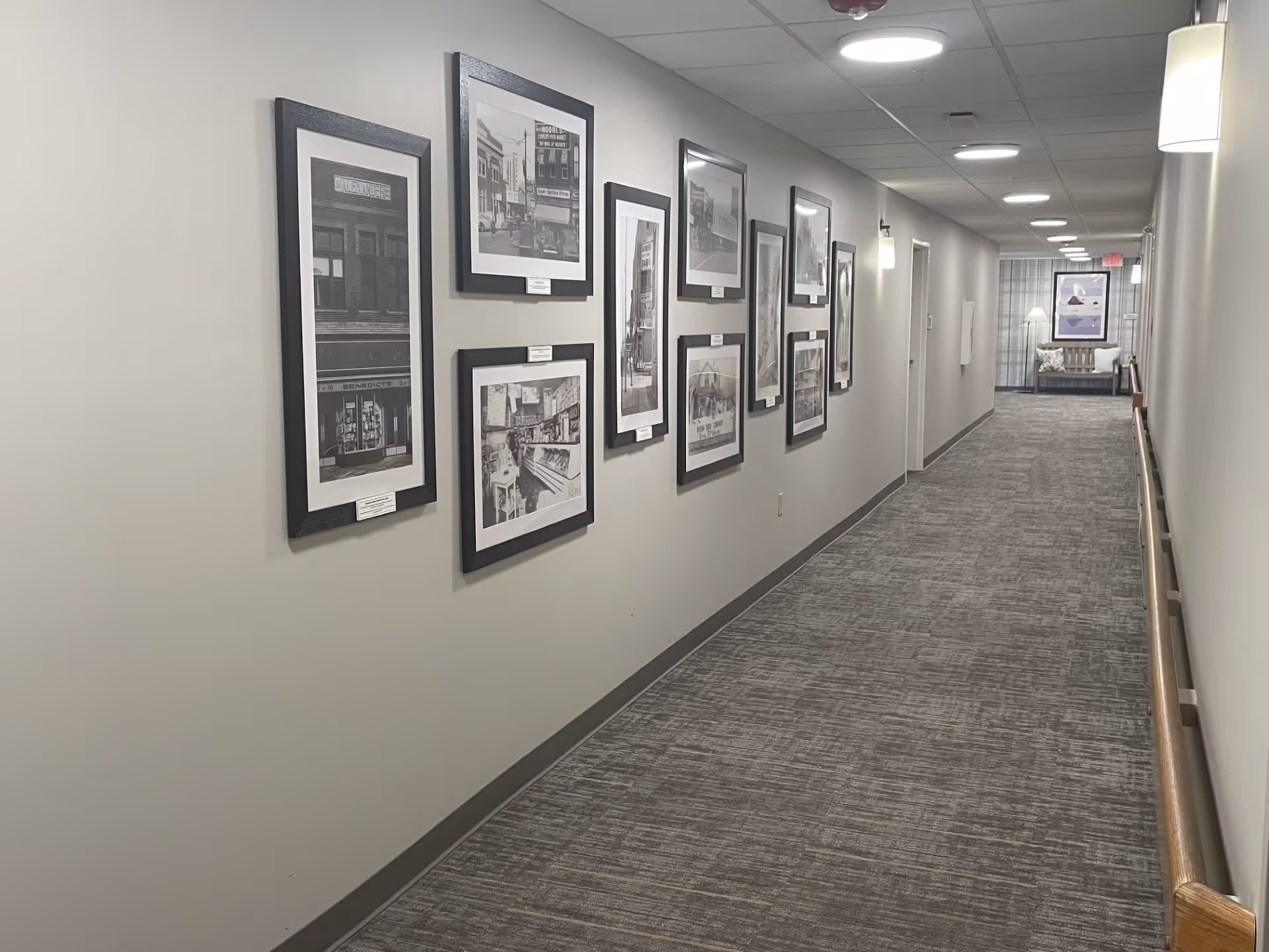 A long, carpeted hallway in a senior living facility with framed black and white photographs on the left wall. The hallway is well-lit with ceiling lights and wall sconces, and has handrails on the right side. At the end of the hallway, there is a seating area with a chair and a framed picture on the wall.