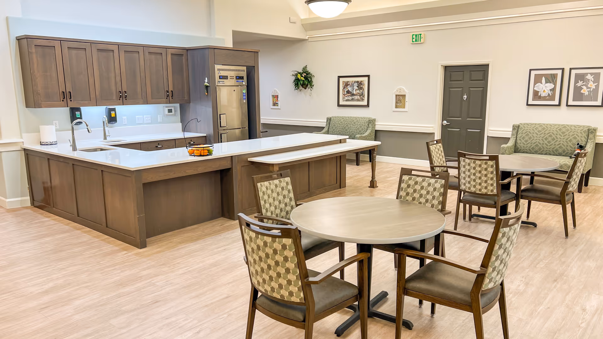 Bright communal dining and kitchenette area with round tables and chairs, a large island, cabinetry, and seating benches.