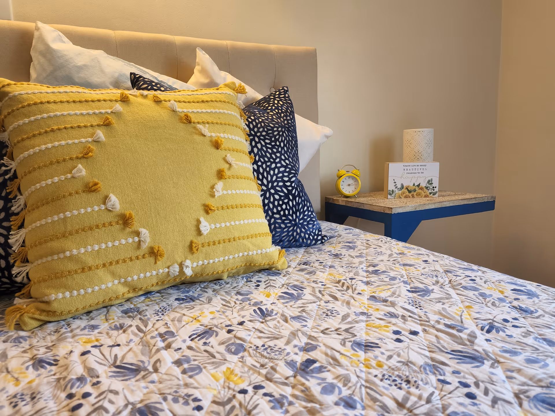 A close-up view of a bed with a beige headboard, decorated with multiple pillows including a large yellow pillow with tassels and smaller blue and white patterned pillows. The bedspread features a floral pattern in shades of blue and yellow. Next to the bed is a small blue floating nightstand holding a yellow alarm clock, a decorative candle holder, and a small framed sign with floral design.