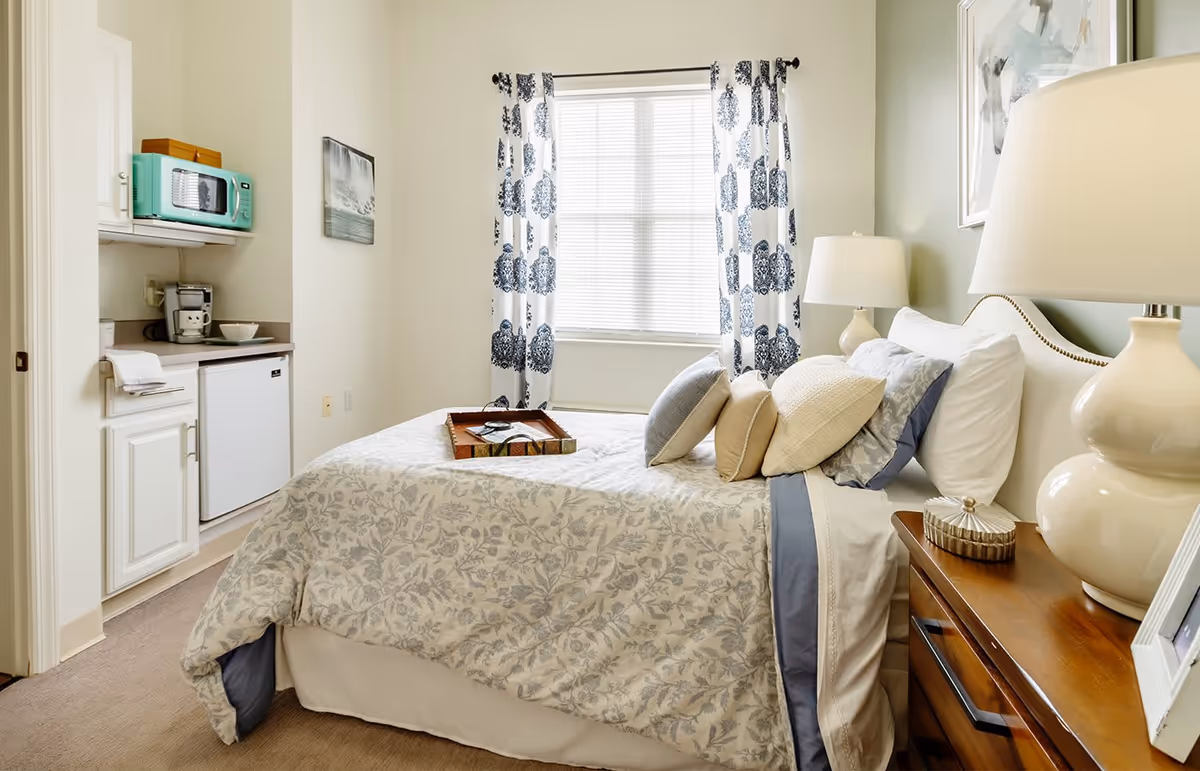 A cozy bedroom with a neatly made bed featuring floral patterned bedding and multiple pillows. There is a wooden nightstand with a lamp and decorative items beside the bed. A window with white blinds and patterned curtains lets in natural light. On the left side, there is a small kitchenette area with a mini fridge, microwave, coffee maker, and cabinets.
